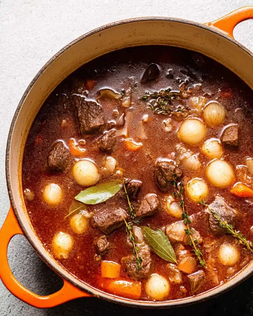 Beef bourguignon simmering with pearl onions, carrots, and herbs in Dutch oven