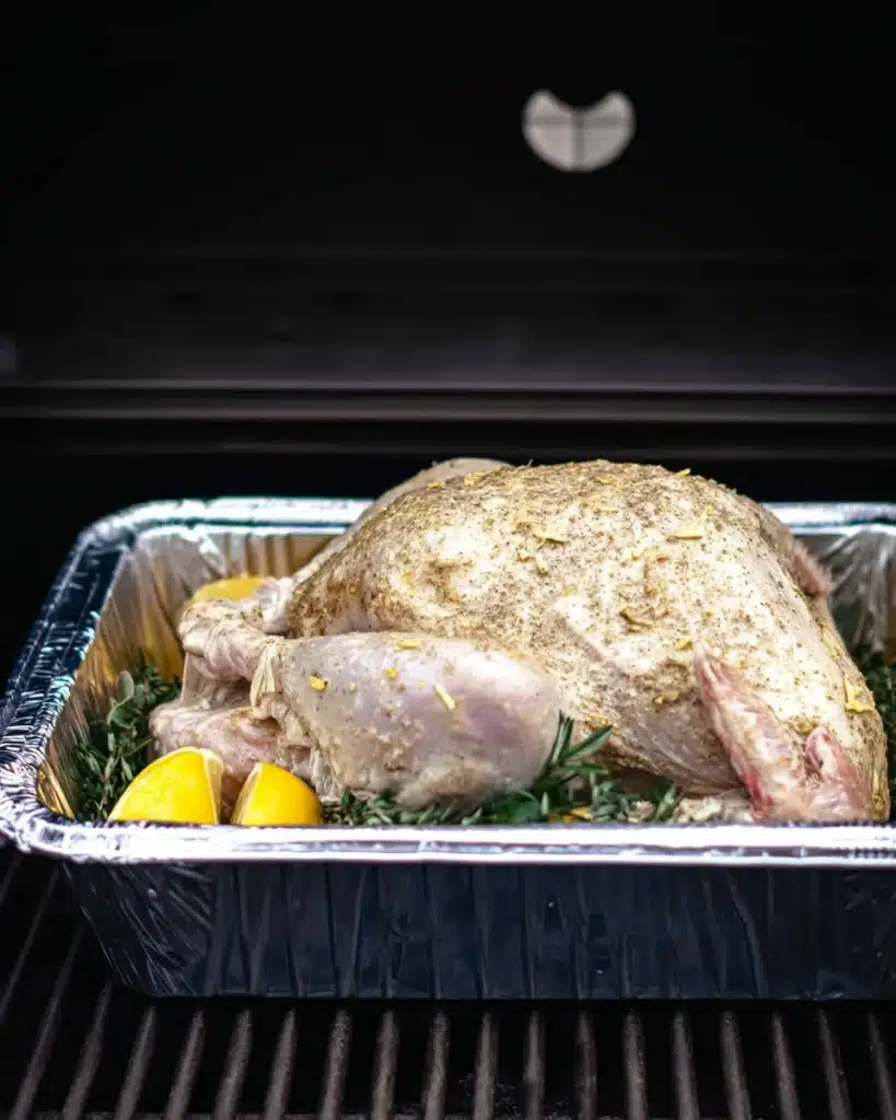 How to Grill Your Turkey on Thanksgiving for Juicy, Crispy Perfection 3 Raw buttered turkey in foil pan placed on grill with lemon and herbs