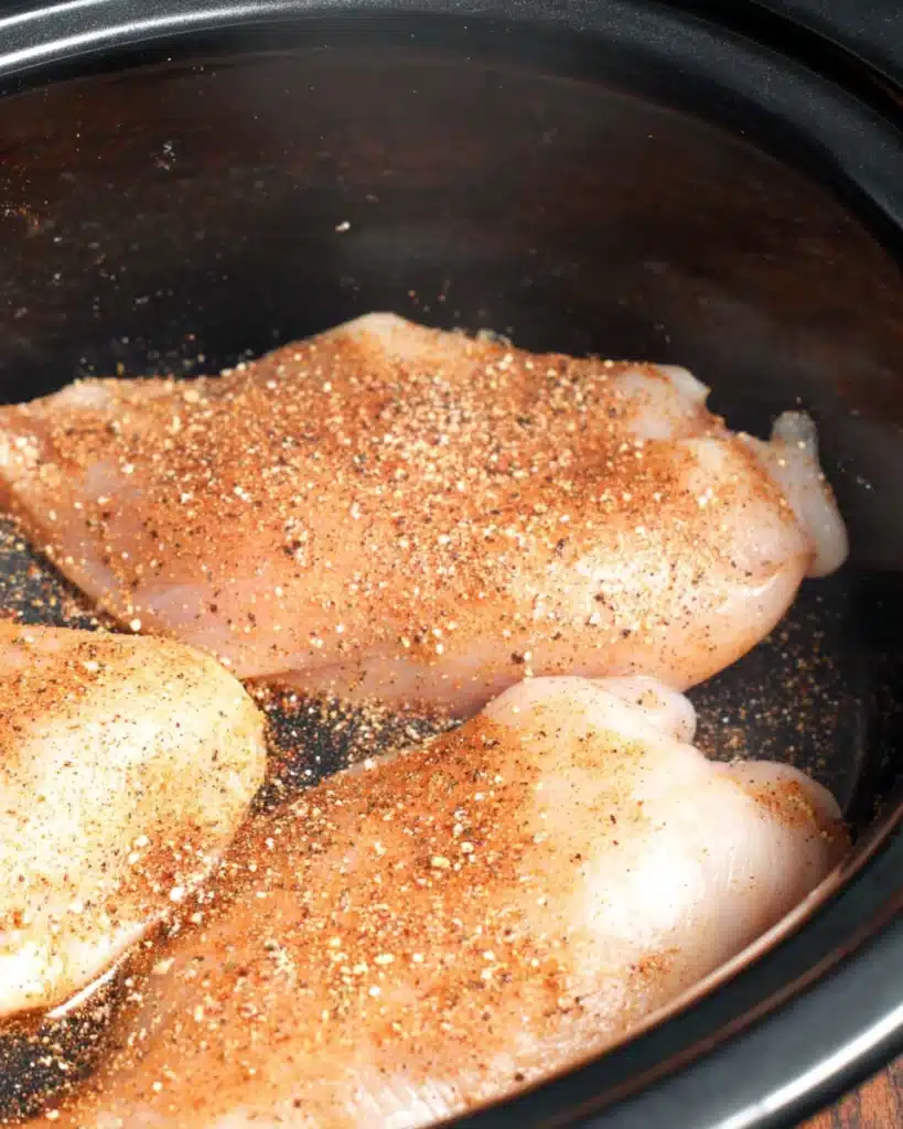 Raw chicken breasts seasoned with taco mix in a slow cooker for chicken enchilada casserole