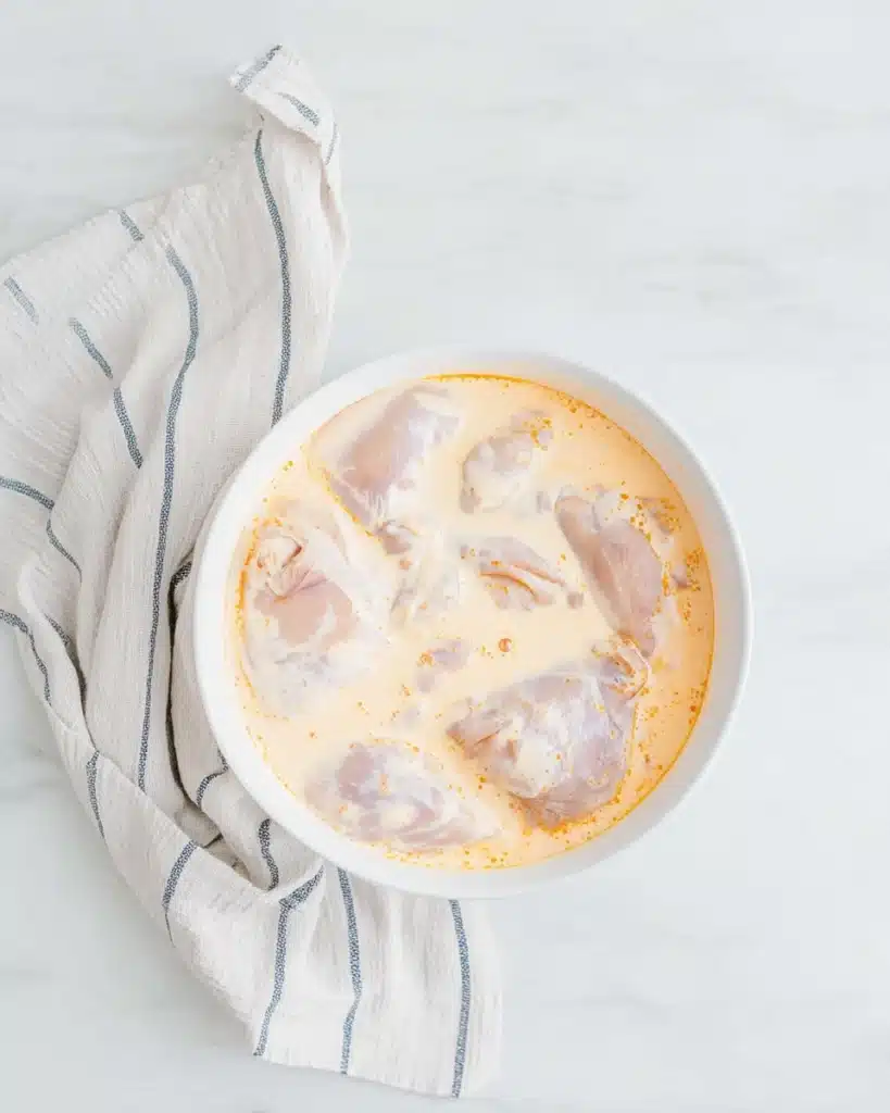 Chicken marinating in creamy buttermilk and hot sauce mixture in a white bowl