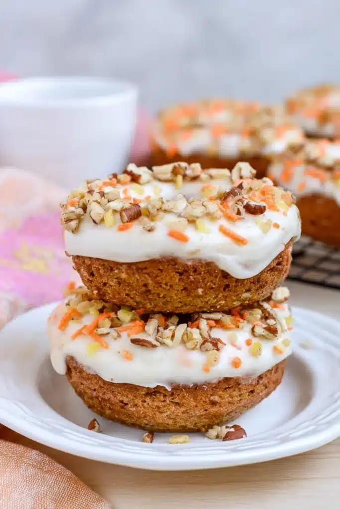 Stacked carrot cake donuts with cream cheese frosting, chopped nuts, and orange sprinkles on a white plate